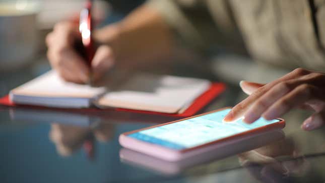 A closeup of a woman's hand tapping a smartphone screen as she writes in a notebook with the other hand