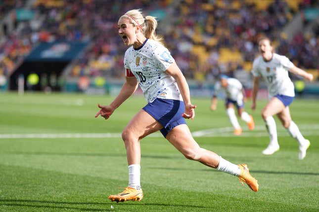 A white women, with blonde hair in ponytail, a pink headband, and a white jersey with blue shorts, yells as she celebrates a goal on a soccer pitch.