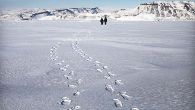 A photo of footprints in the arctic snow with two figures walking in the distance. 