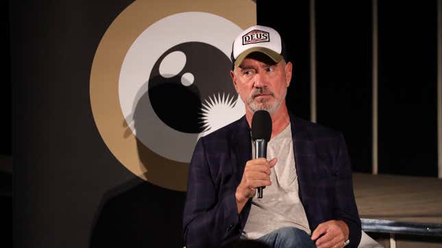 Roland Emmerich, in a white tee, blue jacket, and hat labeled "Deus," sits in front of a backboard with a stylized camera lens logo on it.