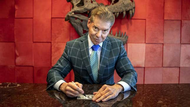 A T-Rex skull mounted against a red wall has its mouth agape behind Vince McMahon who sits at a marble table with a dollar bill in front of him that he is marking with a sharpie as he looks up towards the camera.