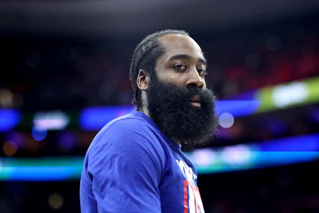A Black man with a bushy beard and his hair in braids, wearing a blue Philadelphia 76ers warm up t-shirt, looks to a camera over his shoulder.