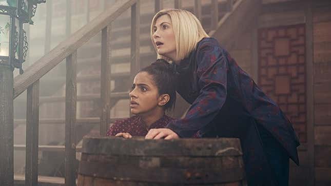 Mandip Gill and Jodie Whittaker as Yaz and the 13th Doctor, hiding behind a barrel as they stare in fear at something off-camera.