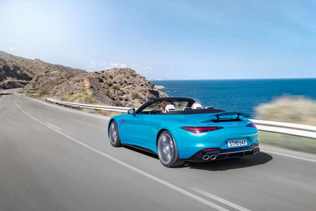 A blue 2023 Mercedes AMG SL43 drives along the coast with its top down and spoiler up, rear three-quarter view