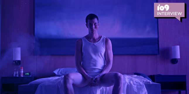 A man wearing an undershirt sits at the edge of a bed in a blue-tinged room.