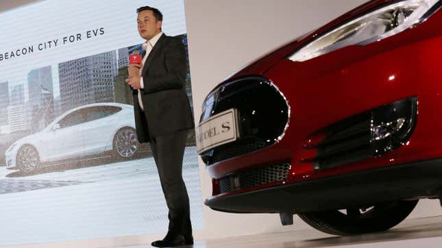 Elon Musk speaking with a red microphone in his hand in front of a red Tesla Model S. In the background is a projection which reads "Beacon City for EVs" and shows a Tesla parked outside of a building.