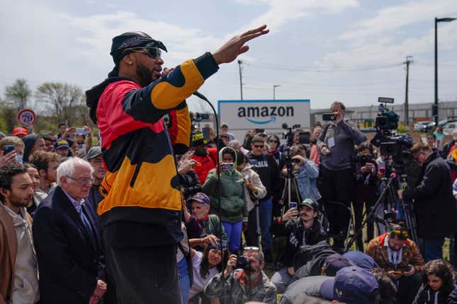 Christian Smalls speaks into a microphone with arm raised amid a crowd of people in front of a large Amazon sign.