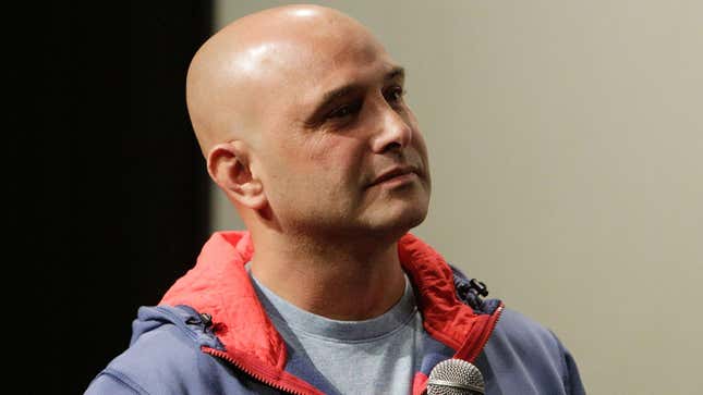 A bald white man in a blue jacket with a red hood listens while holding a microphone.