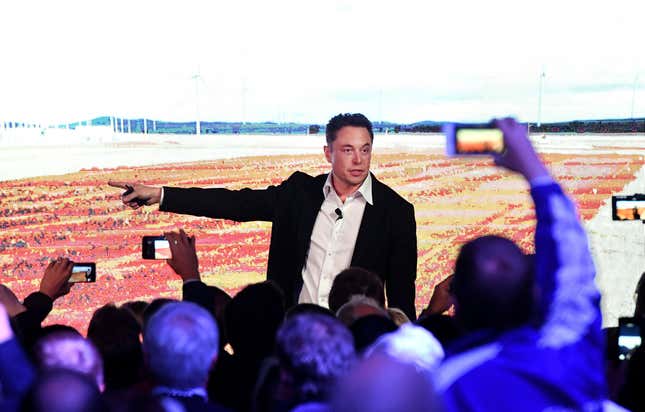 Elon Musk stands in front of a screen showing a highly pixilated empty landscape. People in the crowd hold cell phones up to record his remarks in the foreground. 