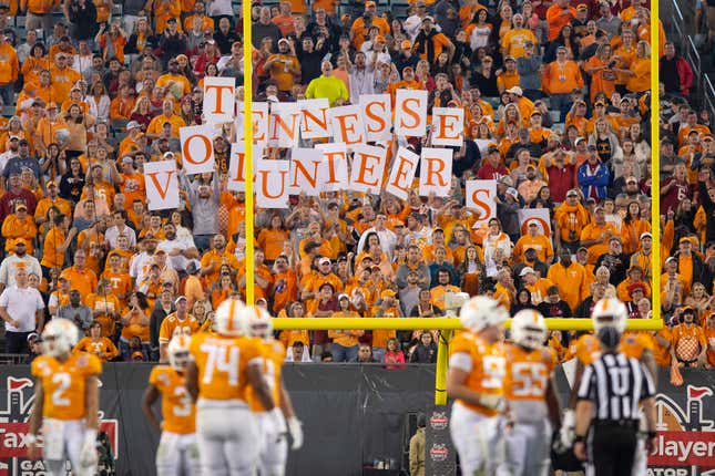 Tennessee Volunteers football fans are seen behind the goal post, most wearing orange, holding up individual letters that spell out Tennessee Volunteers.