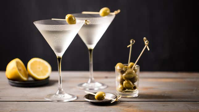 Two martinis sit on a table with a cup of olives, and a lemon cut in half.