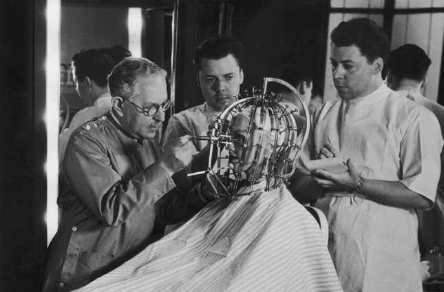 Hollywood cosmetics expert Max Factor (1877 - 1938, left) takes precise measurements of a young woman’s head and face with a ‘Beauty Calibrator’, device, circa 1932.