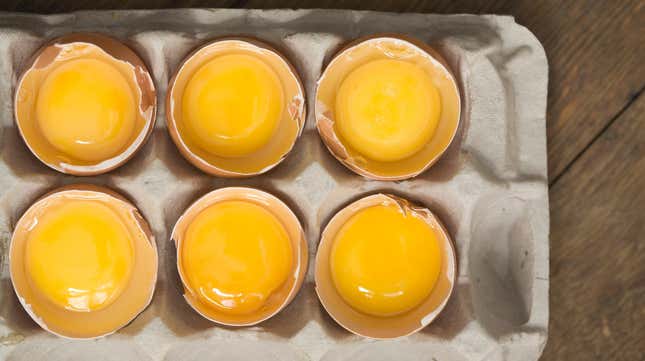 Aerial view of egg carton with each raw egg's top shell removed to show yolk inside