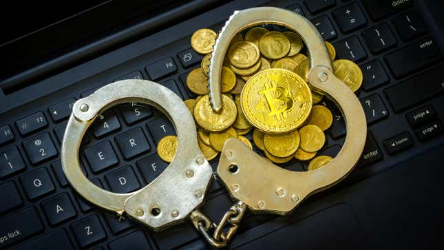 A pair of handcuffs with one cuff open on top of a pile of gold coins bearing the bitcoin logo. This all sits on top of a laptop keyboard.