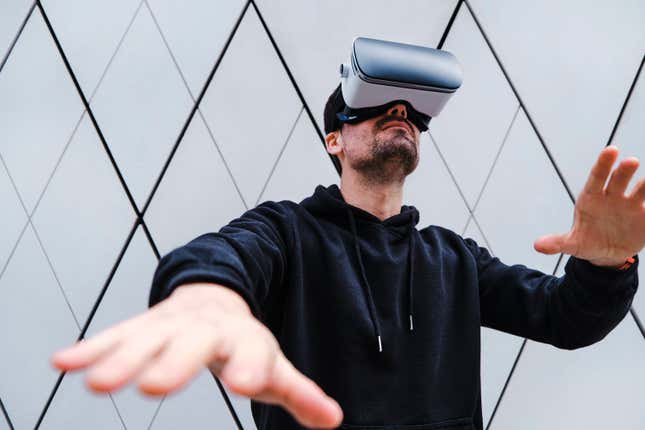 A person in a dark hoodie is wearing a virtual reality headset with their arms out. 