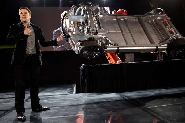 Elon Musk wears a black suit and holds a black mic as he speaks in front of the "skateboard" portion of one of the Tesla vehicles