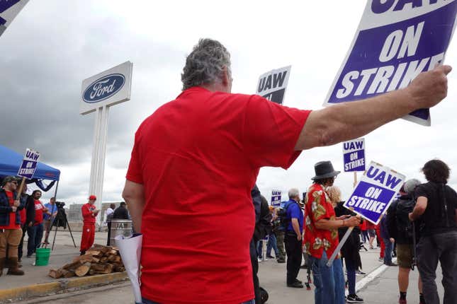 People stand around holding UAW ON STRIKE signs under a cloudy sky outside of a Ford Motor Company plant