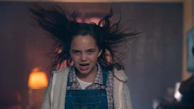A little girl wearing overalls makes an angry face while her hair stands on end.