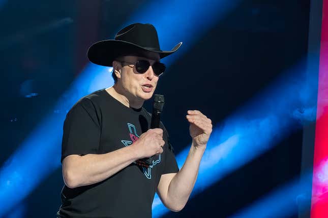 Elon Musk wears sunglasses and a cowboy hat and speaks into a microphone.