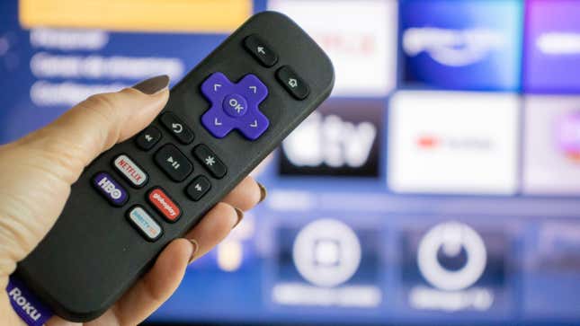 A woman's hand holds a small Roku remote in from of a TV displaying the Roku home screen