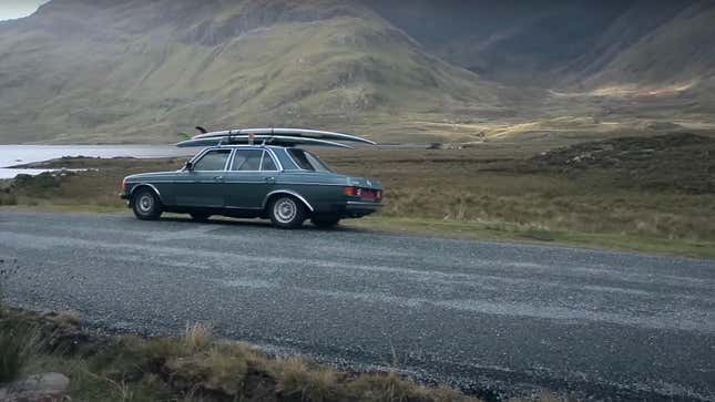 A restored 1980s Mercedes sedan with surfboards on the roof is parked in a mountainous area near water