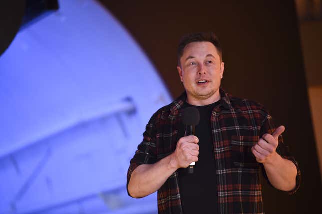 Elon Musk wearing a plaid shirt holds a microphone while speaking to a crowd. 