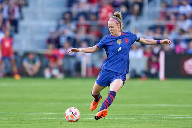 A woman wearing a blue soccer kit with a blonde ponytail, pink headband, and orange soccer cleats kicks an orange and white ball in the center of a green field.