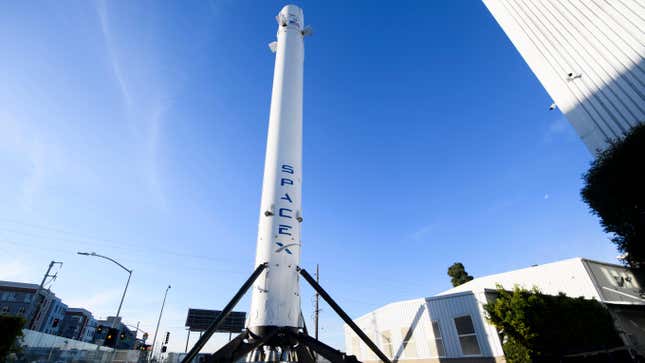 A part of the Falcon 9 Rocket with the SpaceX logo stands up on a large tripod surrounded by nearby buildings