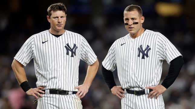 Two white men in white baseball uniforms with blue pinstripes stand with their hands on their hips during an MLB baseball game. 