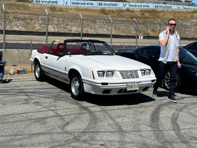 A white convertible 1980s Ford Mustang GT-350 is parked, top down, in the Bring A Trailer lot at Laguna Seca