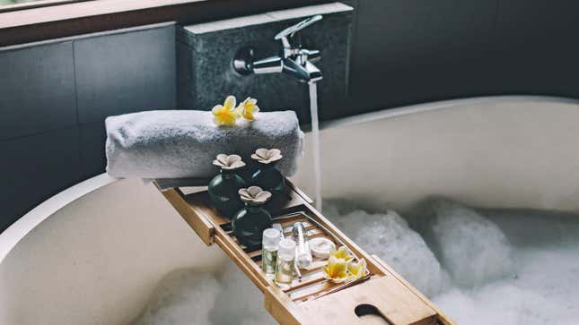 Side-view photograph of a wooden bath tray holding small vials, bottles, a rolled-up towel, and some yellow flower petals. It sits on a white bathtub that's filling up with water and bubbles.