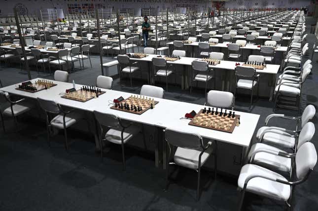 Dozens of tables sporting chess boards and chess clocks in a large room full of empty seats.
