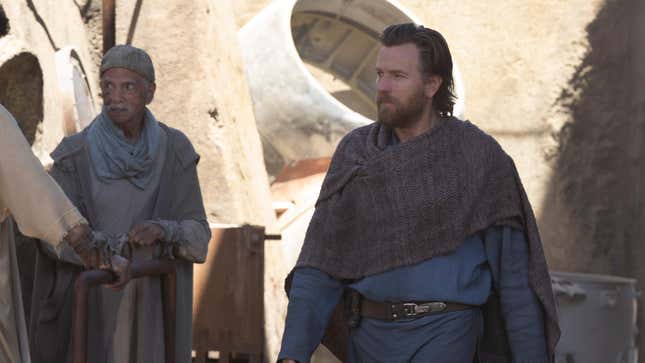 Obi-Wan Kenobi, wearing a poncho and blue robes, walks through a town on Tatooine.