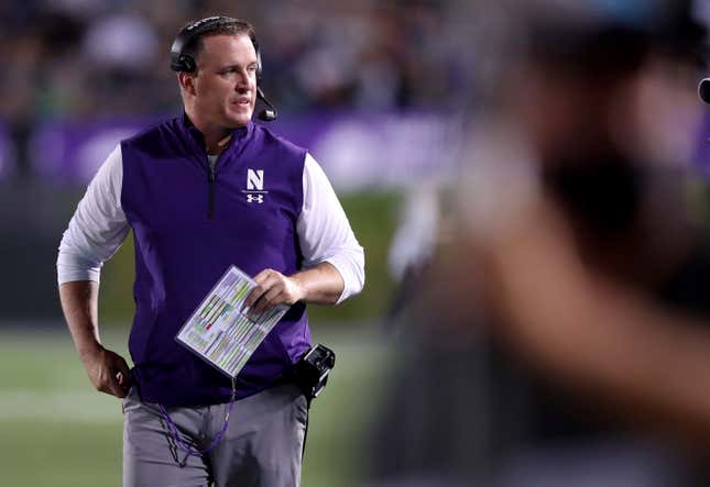 A white man in a white shirt, gray pants, and purple vest wears a headset and carries a play-calling card on the sidelines of an NCAA football game.
