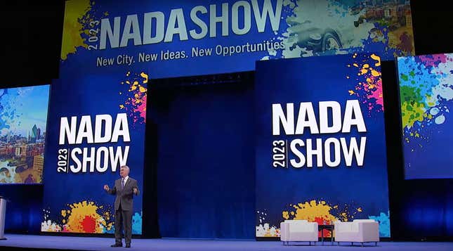 2023 NADA Chairman Geoff Pohanka stands in front of a huge display to give the keynote address for the 2023 NADA Show. The display is blue, and looks splattered with multiple colors. The banner says 20223 NADA SHOW: New City. New Ideas. New Opportunities. 