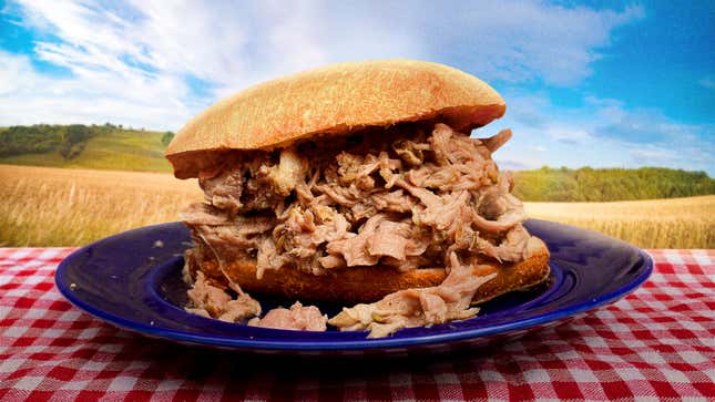 Piled high pork sandwich on blue plate atop a red checkered tablecloth, with a background of fields and blue skies