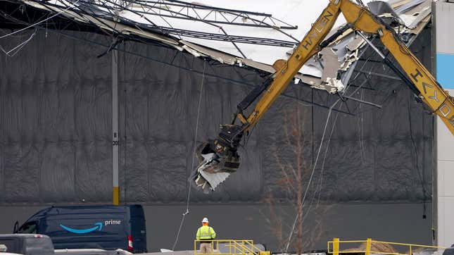 Workers use equipment to remove a section of roof left on a heavily damaged Amazon fulfillment center in Edwardsville, Illinois. An Amazon Prime delivery van sits in the lower left corner.