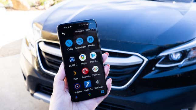 A photo of a hand holding up a Pixel 5 with the Assistant Driving Mode interface on it, with a car in the background