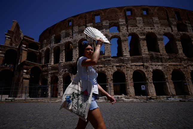A woman with a tote bag walks past the Roman Colosseum holding up a fan in front of her face to block out the sun.