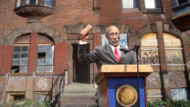 Mayor David Bing christens the brand-new housing development by shattering the window of a dilapidated tenement building.