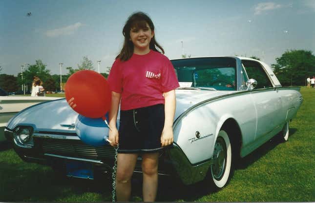 A girl in a pink shirt and black shorts holds a red balloon in front of a classic Ford Thunderbird