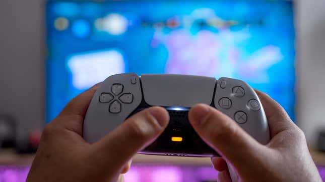Someone holds the PlayStation 5's DualSense controller in front of a blurred TV screen.