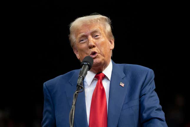 Donald Trump, in a blue suit, red tie, and white shirt, speaks into a microphone in front of a black background