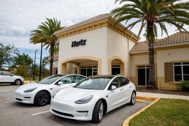 Two white Tesla Model 3s parked outside a Hertz rental office.