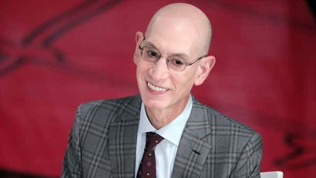 A bald, smiling white man wears a plaid suit and tie and smiles before a red background