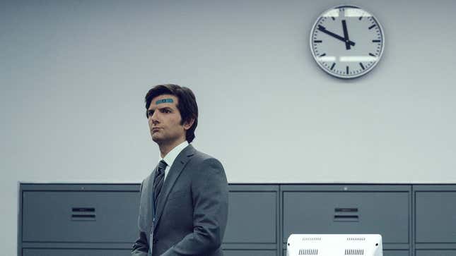 Adam Scott sits on a desk wearing a gray suit with a clock behind him