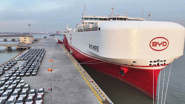 Luftaufnahme von Reihen von Elektrofahrzeugen auf einem Dock neben einem großen weiß-roten Schiff im Wasser neben dem Hafen