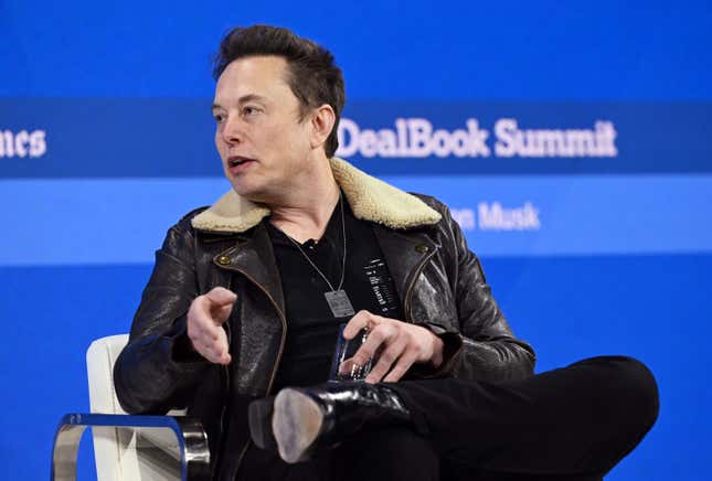 Elon Musk sitting in a chair in front of a blue backdrop that says DealBook Summit