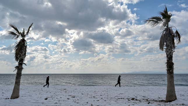 People walk along a snow-covered beach between two palm trees in Athens on January 25, 2022