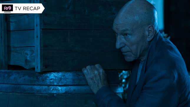 Jean-Luc Picard, in the dark, hides behind a stack of barrels and boxes, resting one hand on the pile.